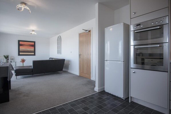 A modern kitchen with a refrigerator, oven, and a dining area with a sofa and table. - Maritime Quarter - 2 Bedroom Apartment - SA1 Beach Front (Fisherman's Way) (Swansea)