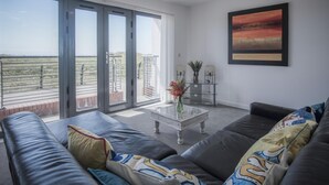 A modern living room with a large sliding glass door leading to a balcony, a glass coffee table, a dark leather sofa, and a framed artwork on the wall. - Maritime Quarter  - 2 Bedroom Apartment - SA1 Beach Front (Fisherman's Way) (Swansea)