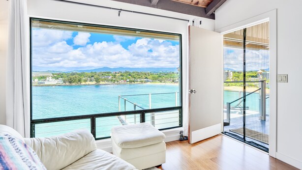 A room with a large window overlooking a body of water, a balcony with a railing, and a white sofa.