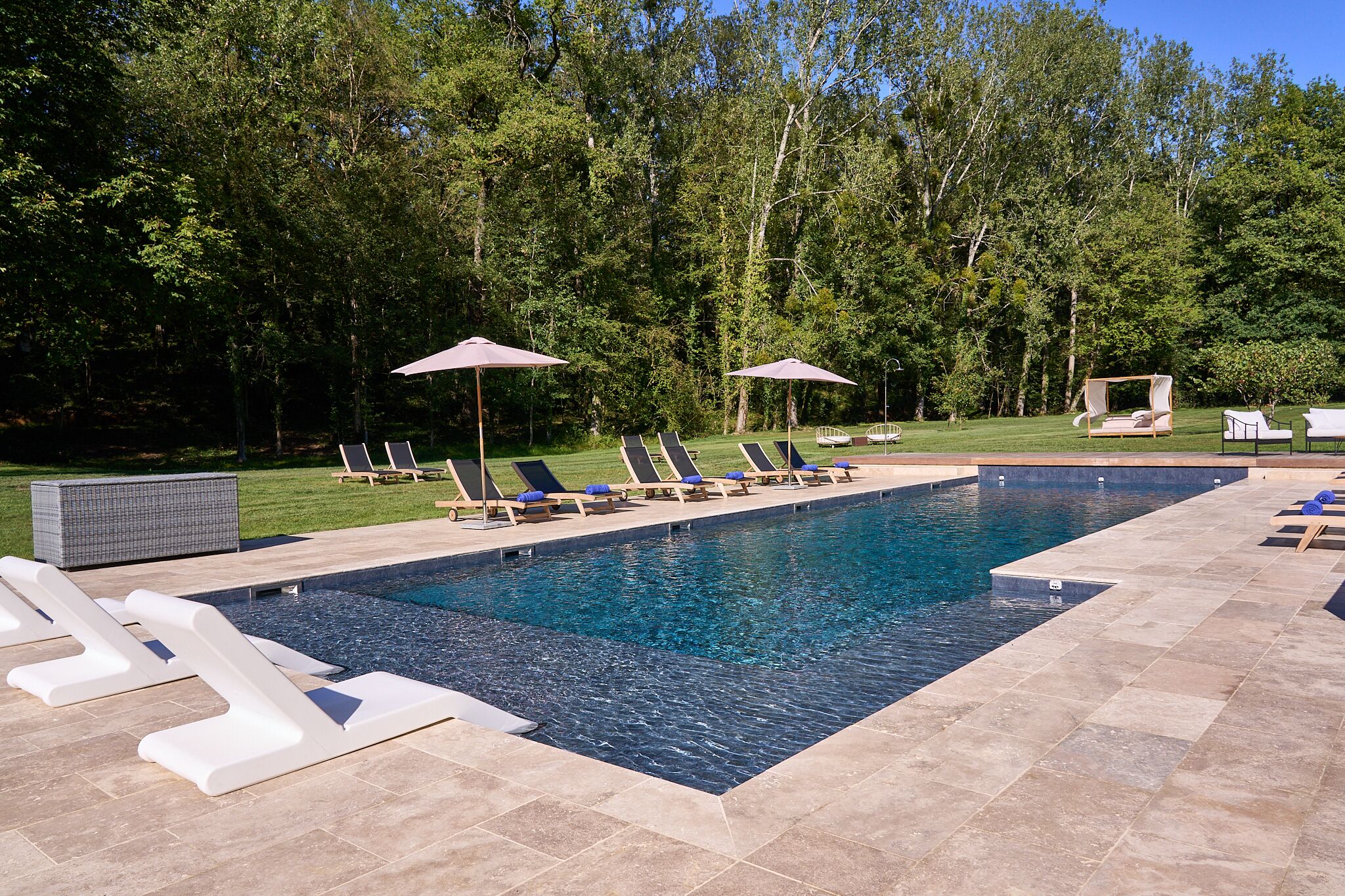 A rectangular pool with clear blue water, surrounded by white lounge chairs and umbrellas, set against a backdrop of dense trees.