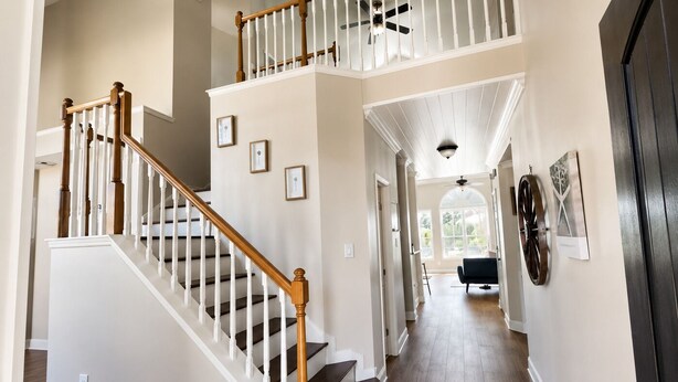 Un interior de varios niveles con una escalera de madera, un pasillo que conduce a una habitación con ventilador de techo y una puerta con pomo decorativo.