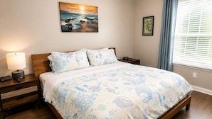 Un dormitorio con una cama de madera, una mesita de noche con lámpara, un cuadro en la pared y una ventana con cortinas.