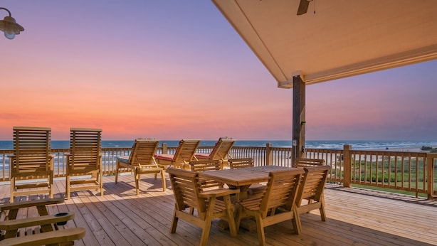 Une terrasse en bois avec des chaises longues et une table, offrant une vue sur l’océan au coucher du soleil.