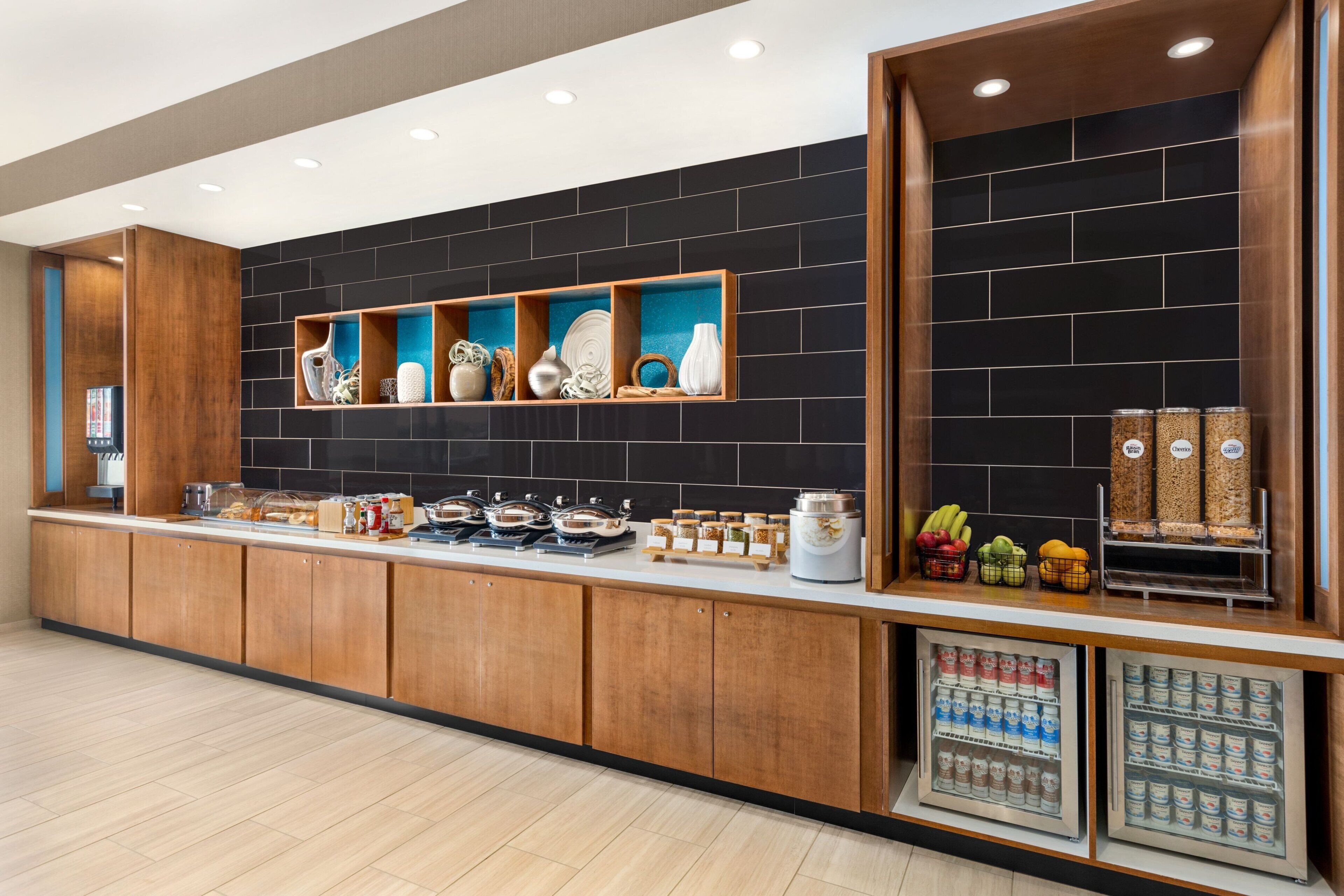 A modern kitchen with wooden cabinets, a black tiled backsplash, and various food items displayed on shelves and counters.