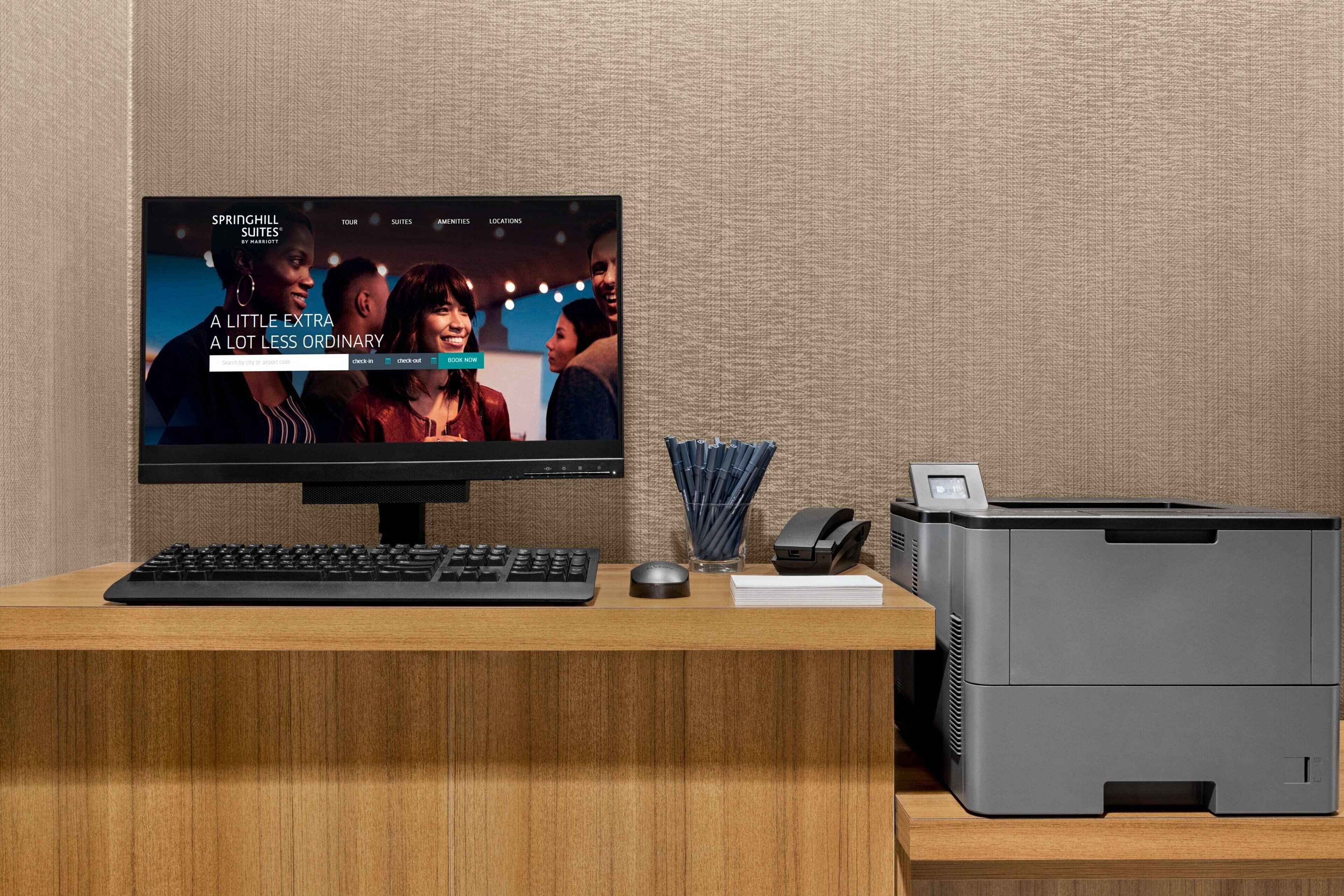 A hotel reception desk with a computer monitor displaying a webpage, a keyboard, a mouse, a printer, and a stack of papers.