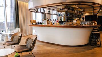 A modern bar area with a curved counter, bar stools, and a variety of bottles and glasses. There are two cushioned chairs and a small table with a plant.