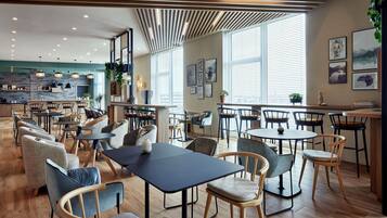 A modern restaurant with wooden flooring, multiple dining areas, and large windows providing natural light.