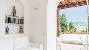 Ein weißes Sofa, ein Bücherregal mit Büchern und Figuren sowie ein Blick auf eine Stadtlandschaft durch einen offenen Durchgang.