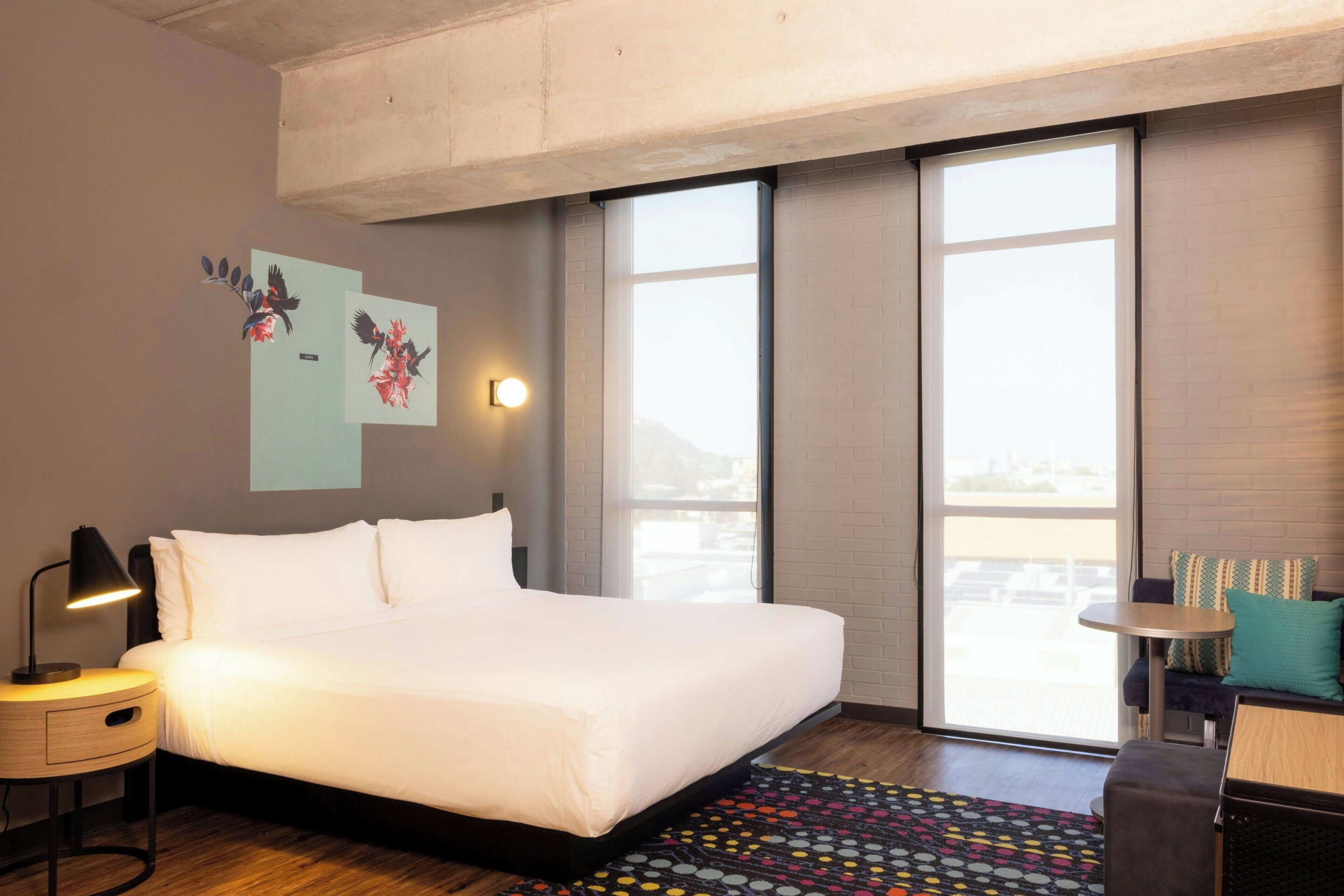 A modern hotel room with a large bed, a small desk, a chair, a sofa, and a large window offering a city view.