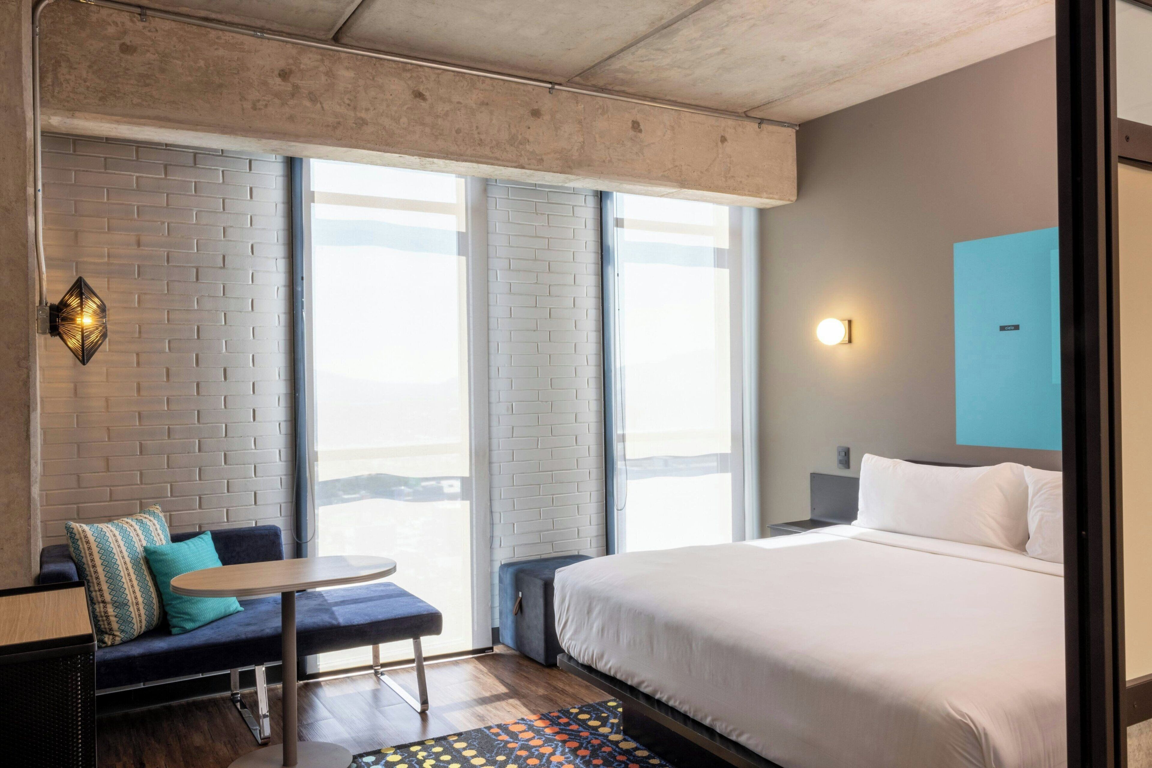 Hotellihuone, jossa on suuri sänky, tyynyillä varustettu sohva, pieni pöytä ja näkymä suurista ikkunoista.
