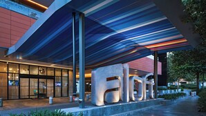 A modern building with a glass facade, large metallic letters spelling "hotel," and a covered walkway with plants.