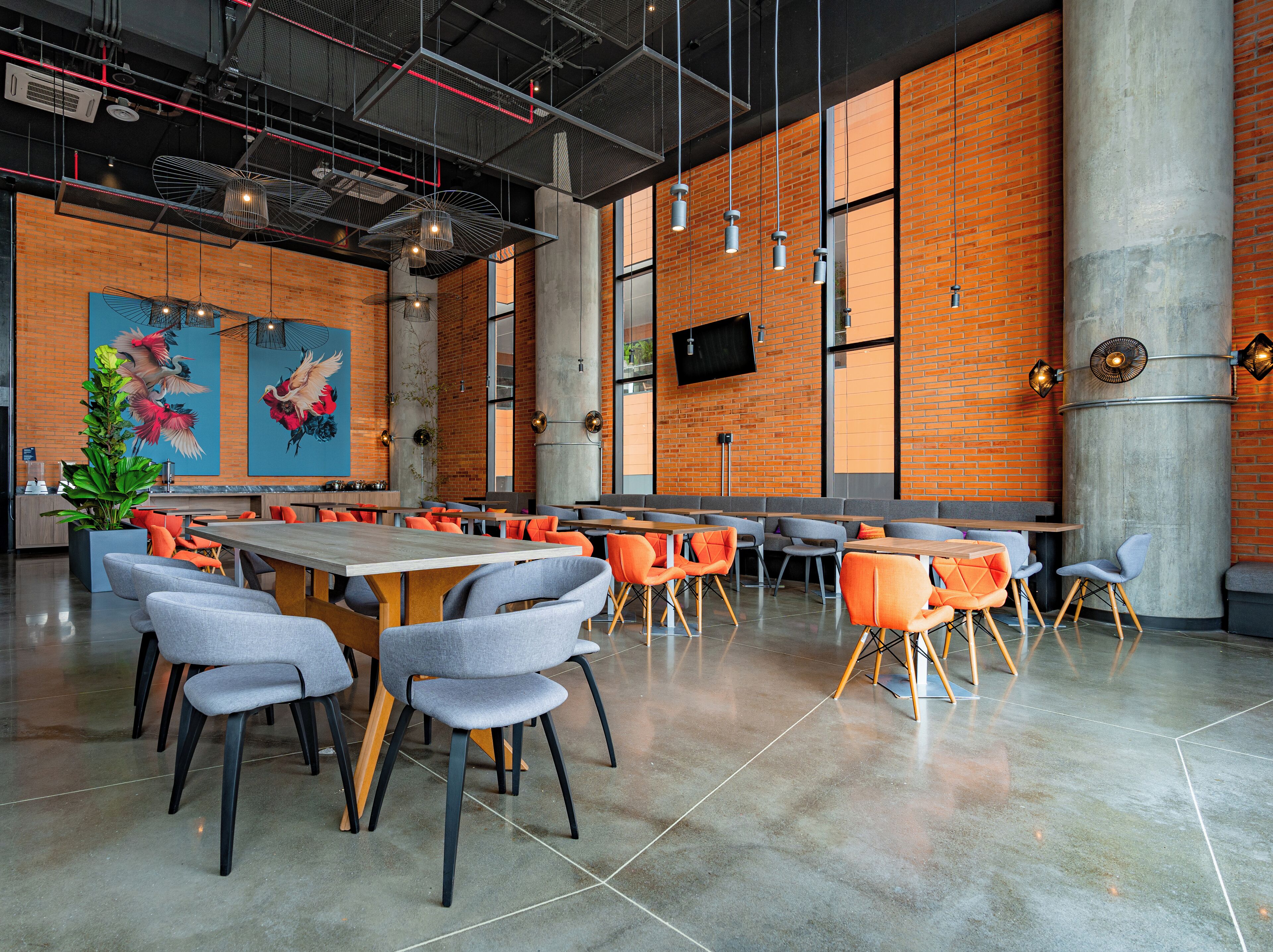A modern dining area with high ceilings, exposed brick walls, and a mix of grey and orange chairs.