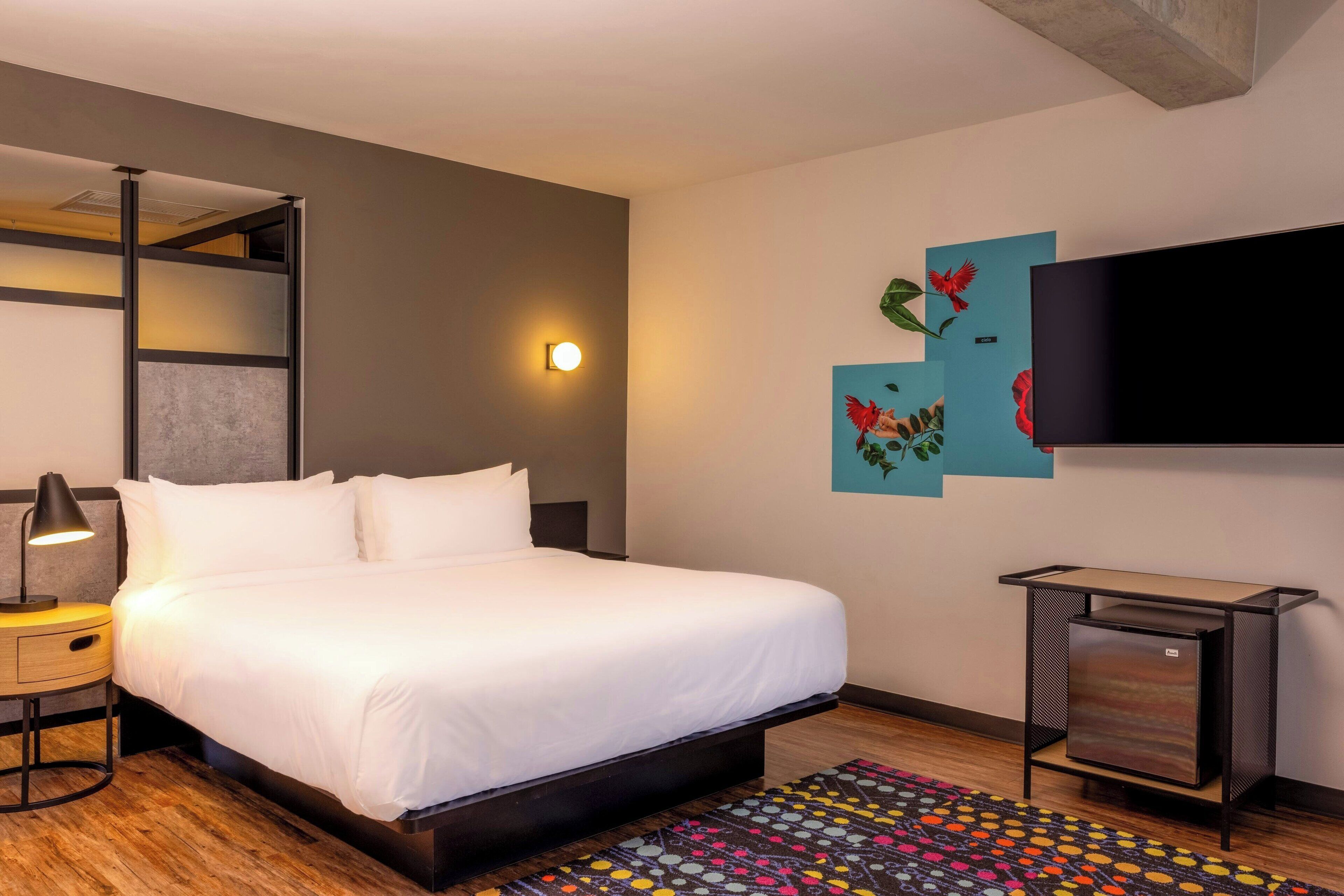 A modern hotel room with a large bed, a bedside lamp, a small table, a TV mounted on the wall, and a colorful rug.
