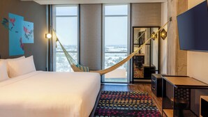 Une chambre d’hôtel avec un grand lit, une vue sur la ville grâce à de grandes fenêtres, un hamac, un bureau et une télévision.