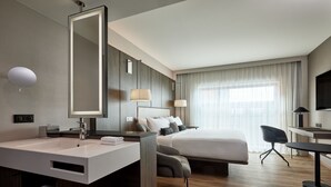Habitación de hotel moderna con una cama grande, un escritorio, una silla, un baño con lavamanos y espejo, y un ventanal con cortinas.