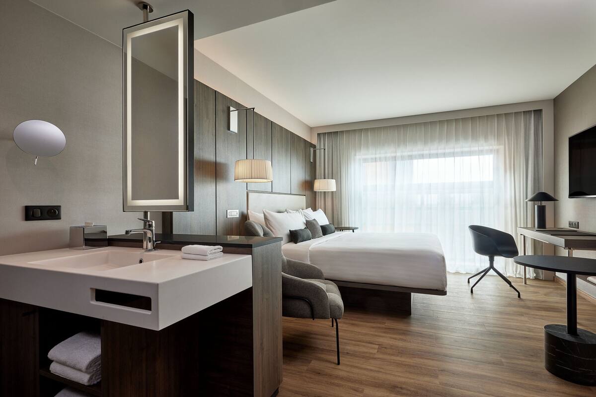 A modern hotel room with a large bed, a desk, a chair, a bathroom with a sink and mirror, and a large window with curtains.