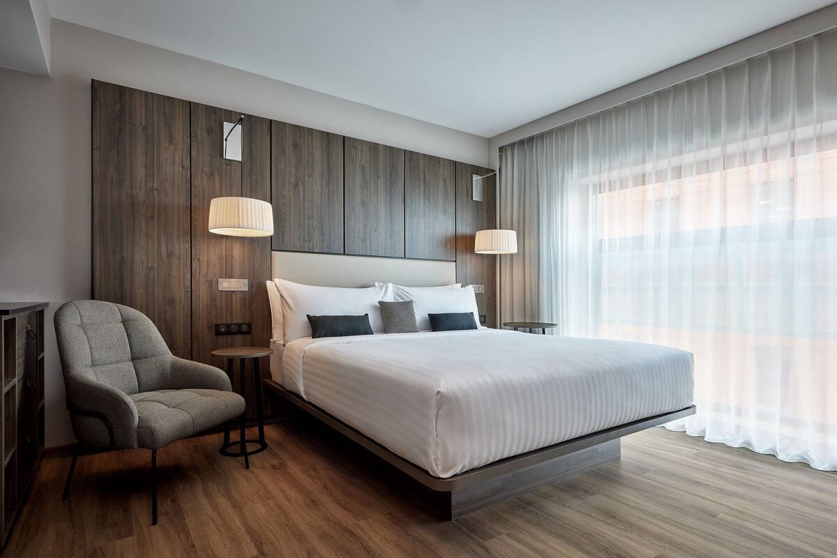 A modern hotel room with a large bed, a grey armchair, wooden panel walls, and a large window with sheer curtains.