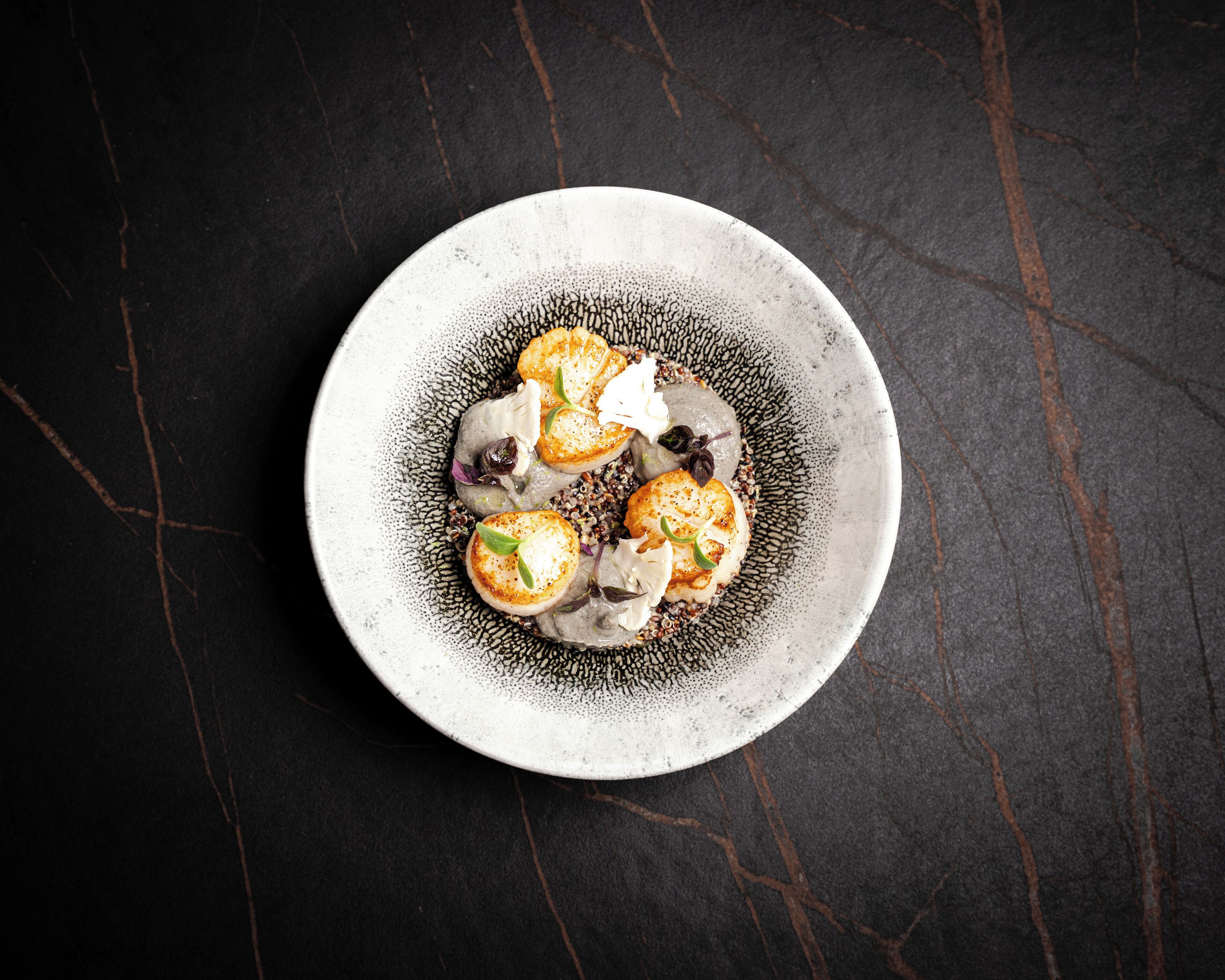 Un plato de cerámica redondo con un diseño texturizado, que contiene varios vieiras, una cucharada de crema y una guarnición, sobre una superficie oscura y agrietada.