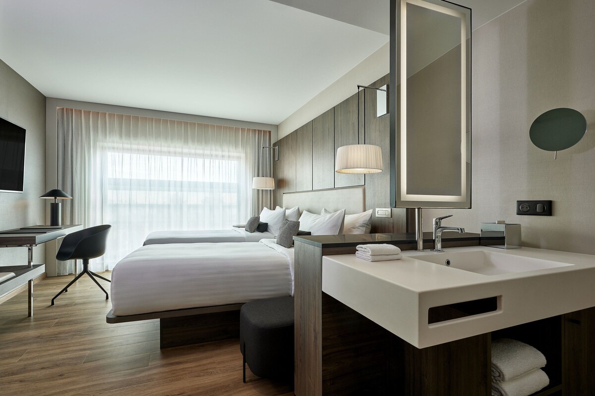 Una habitación de hotel moderna con una cama grande, un escritorio, una silla, un tocador con espejo y un ventanal con cortinas.