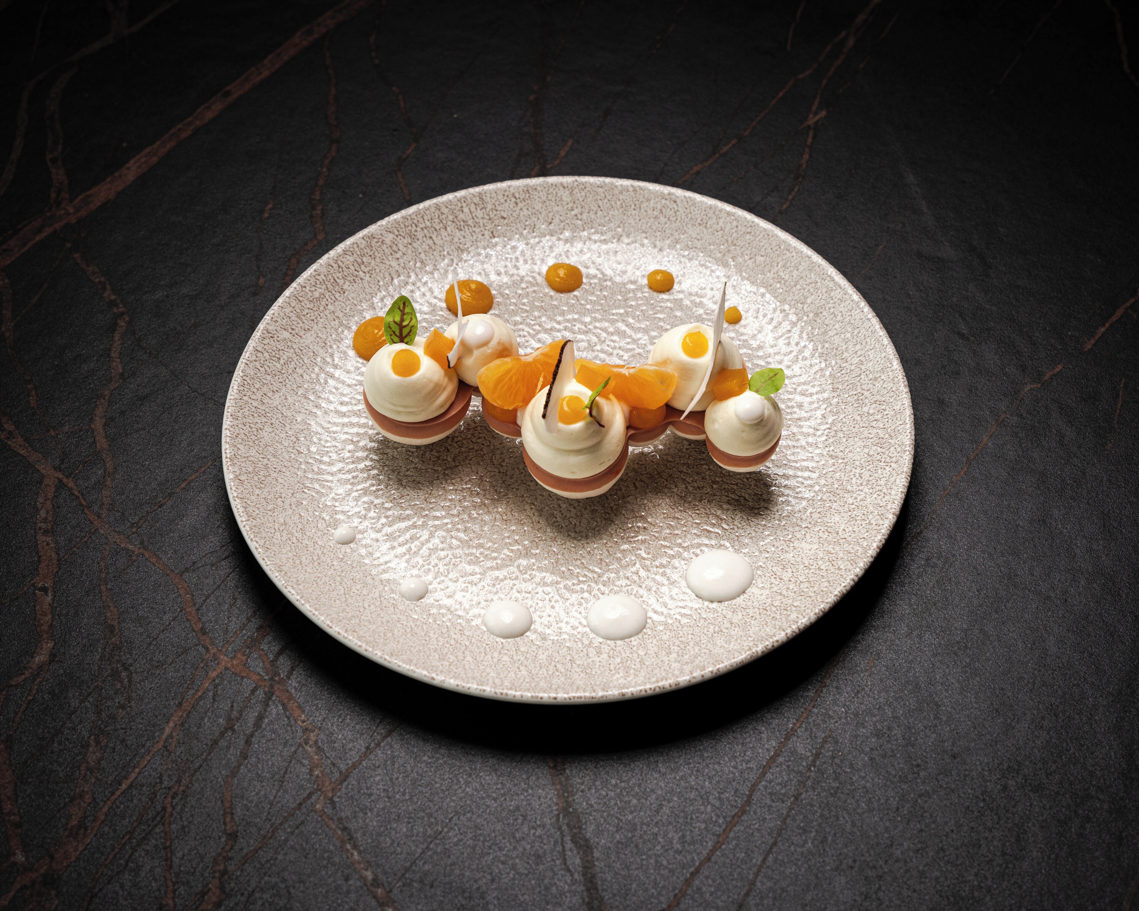 A round plate with a dessert featuring spherical shapes topped with orange slices and a green leaf, set on a dark textured surface.