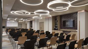 Uma sala de conferências com fileiras de cadeiras, uma tela grande e luminárias circulares modernas no teto.