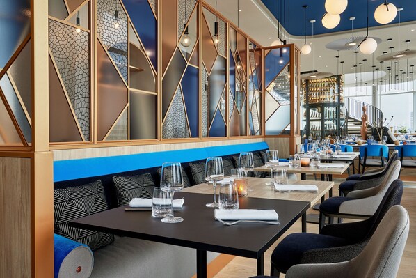 Un restaurante moderno con paneles geométricos en las paredes, asientos azules y mesas ordenadas con vasos y servilletas.