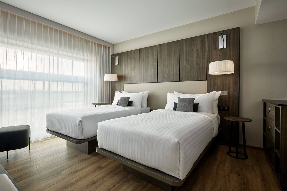 A hotel room with two beds, a wooden headboard, a small table, and a lamp.