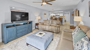 Un salon moderne avec un téléviseur à écran plat, un meuble de rangement bleu, un canapé beige, un pouf à motifs et un ventilateur de plafond.