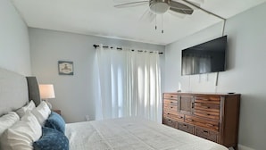 Une chambre à coucher comprenant un lit, une commode, un ventilateur de plafond, un téléviseur et une fenêtre avec des rideaux.