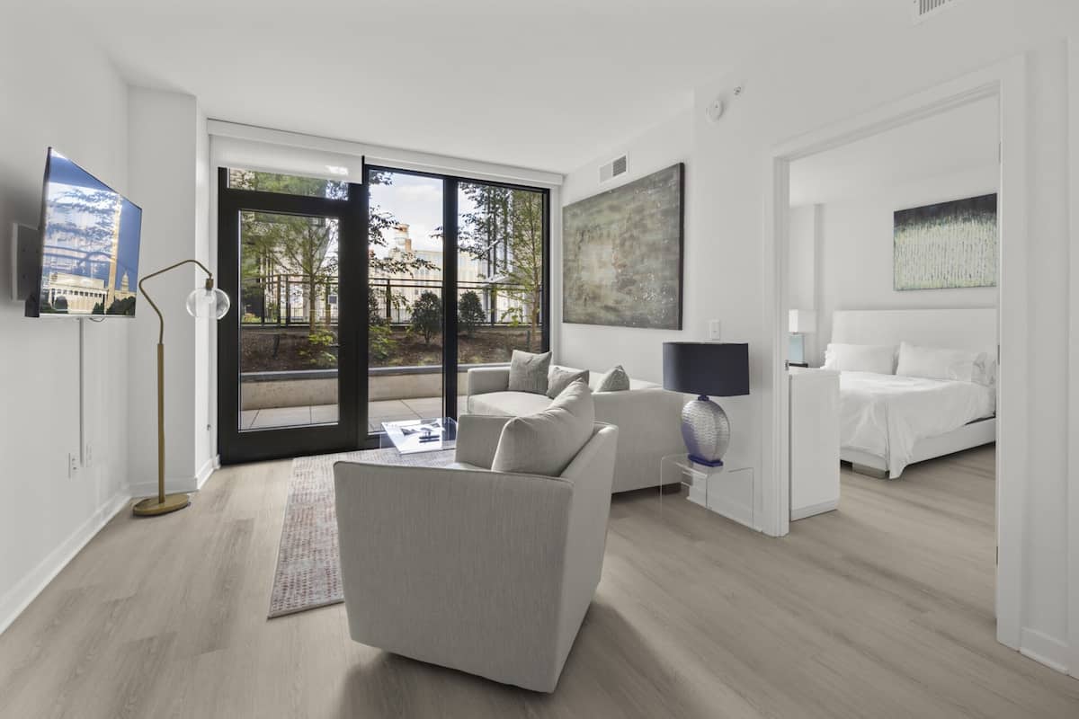 Habitación de hotel moderna con sofá, cama, televisor y un ventanal con vistas a árboles y edificios.