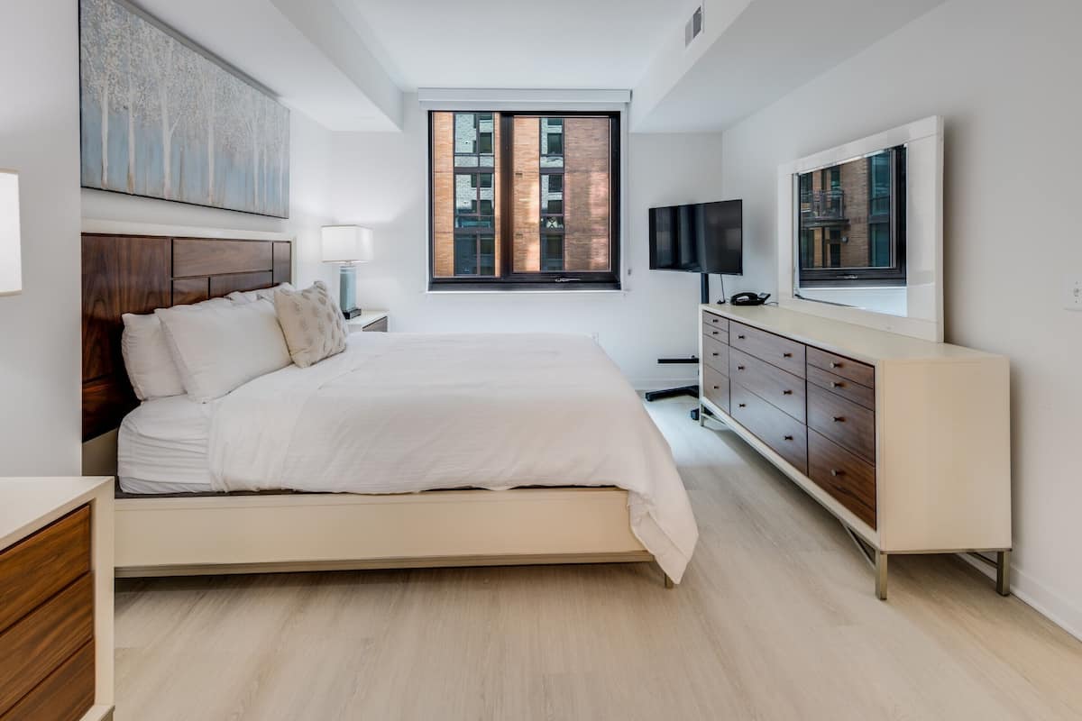 Un dormitorio moderno con cama, cómoda, televisor y ventana con vista a edificios.