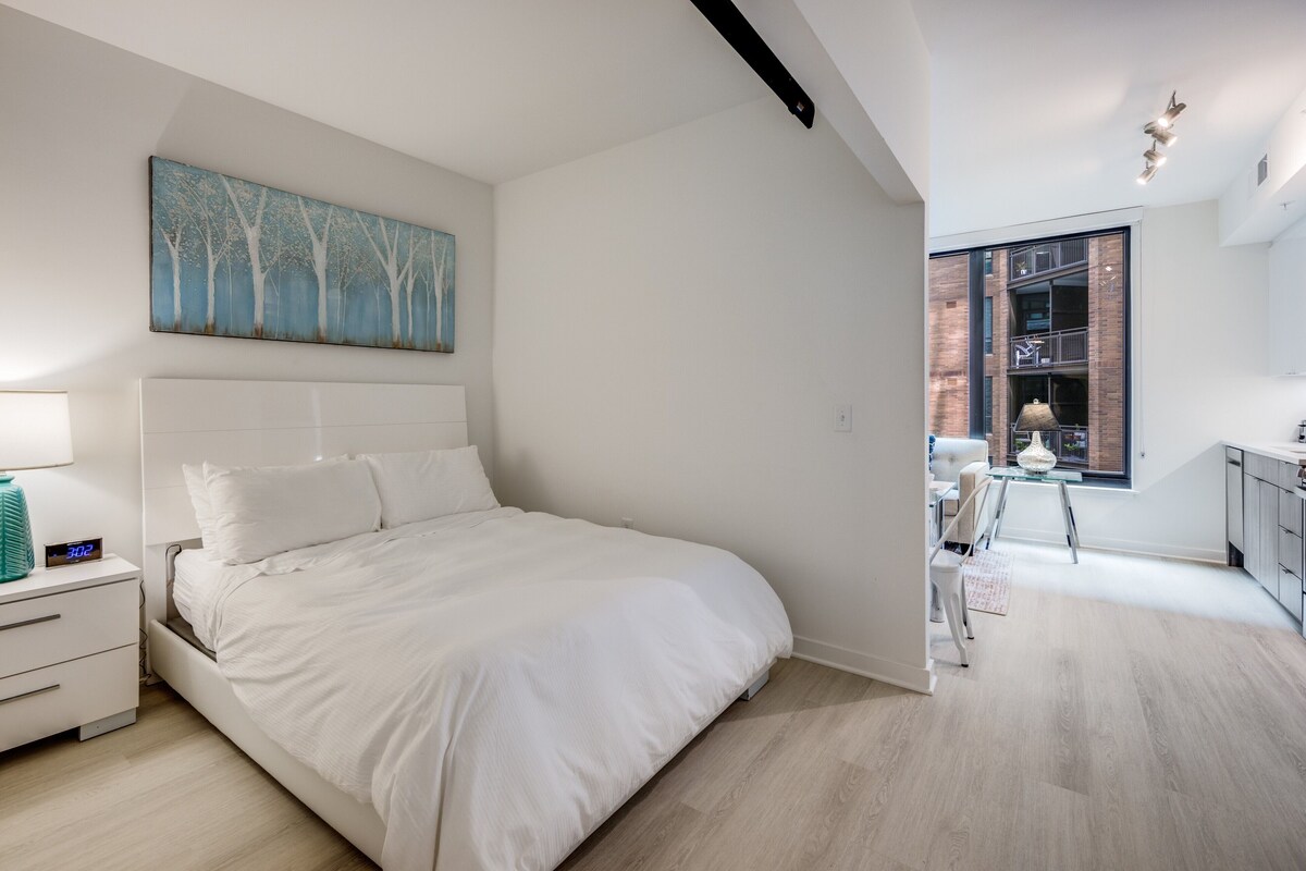 A modern bedroom with a large bed, a nightstand, a painting, and a view of a kitchen and balcony.