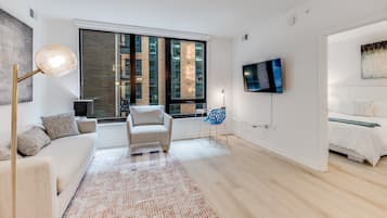 Un salon moderne avec un canapé, un fauteuil, un téléviseur et une grande fenêtre donnant sur un immeuble.