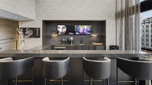 Zona de bar moderna con mostrador de mármol, taburetes negros y una pantalla grande que muestra un reloj y el número de una habitación de hotel.