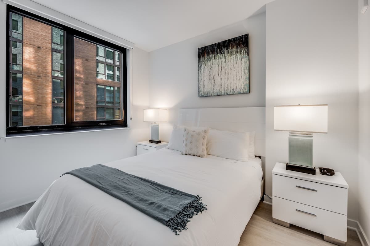 Un dormitorio moderno con una cama grande, lámparas de noche, una mesita de noche, una ventana con vistas a edificios y un cuadro abstracto colgado en la pared.