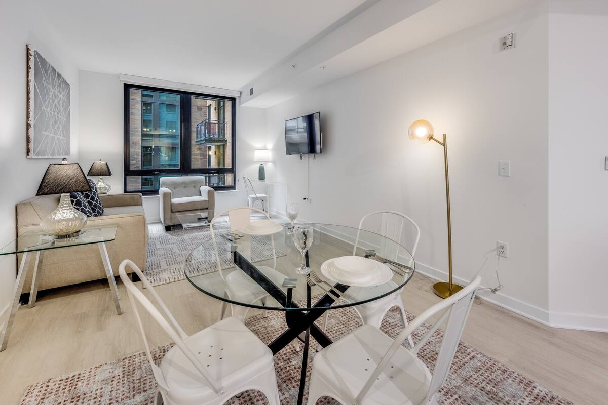 Una sala de estar moderna con una mesa de comedor de vidrio, sillas blancas, un sofá y un televisor montado en la pared.