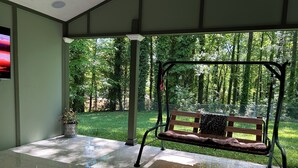 Un espace extérieur abrité, avec un banc et une table, entouré de verdure et d’une clôture.