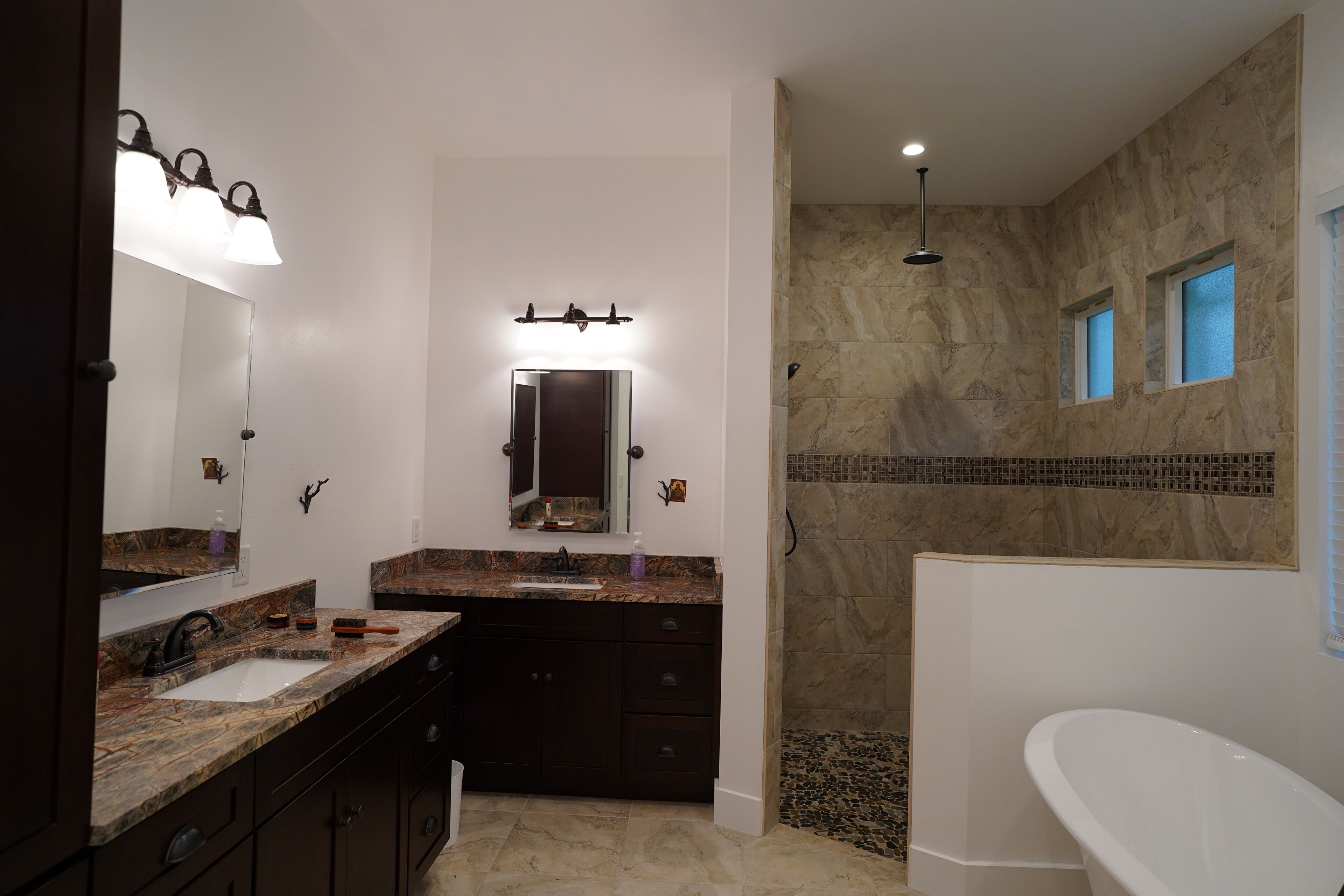 A modern bathroom with a large mirror, dual sinks, a shower with a stone wall, and a freestanding bathtub.