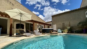 Una piscina con sillones de playa y sombrillas, rodeada por un edificio de techo con tejas y paredes de piedra.