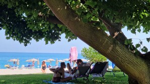 A beach scene with a tree providing shade, a grassy area, and beach chairs and umbrellas in the background. - Nina's house 2 300 meters from the sea (Santa Caterina Dello Ionio Marina)