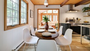 Un coin repas avec une table en bois, des chaises blanches et un vase de fleurs. De grandes fenêtres offrent une vue sur l’extérieur.