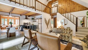Un vaste espace de vie doté d’un plafond haut, de poutres en bois, d’un escalier et d’une cuisine située à l’arrière-plan.