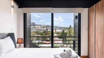 A hotel room with a large window, a bed with white linens, a bedside table with a lamp, and a view of a cityscape.