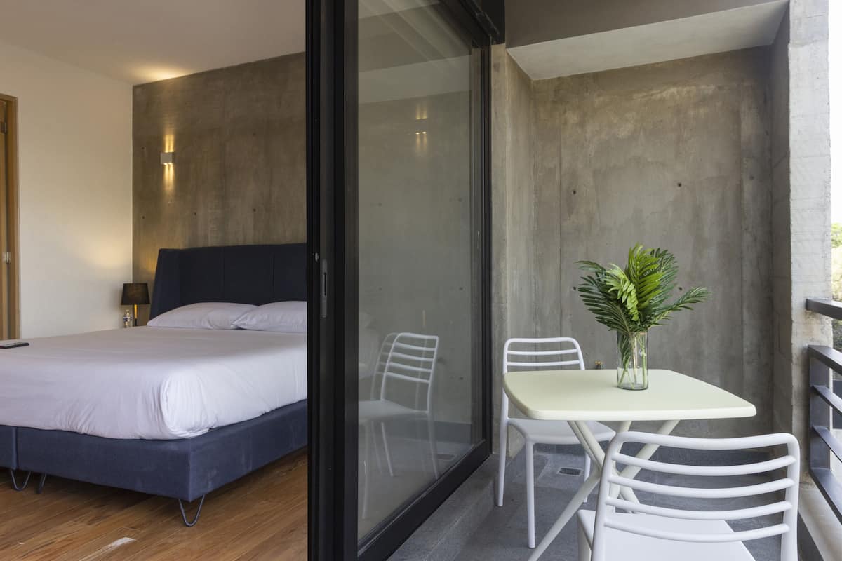 Quarto moderno com uma cama grande, porta de vidro para uma varanda com mesa e cadeiras, e um pequeno vaso de planta.