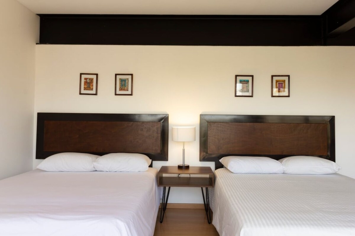 A hotel room with two beds, a nightstand with a lamp, and framed pictures on the wall.