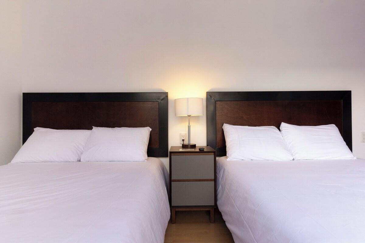 A hotel room with two single beds, a nightstand with a lamp, and a small table between the beds.