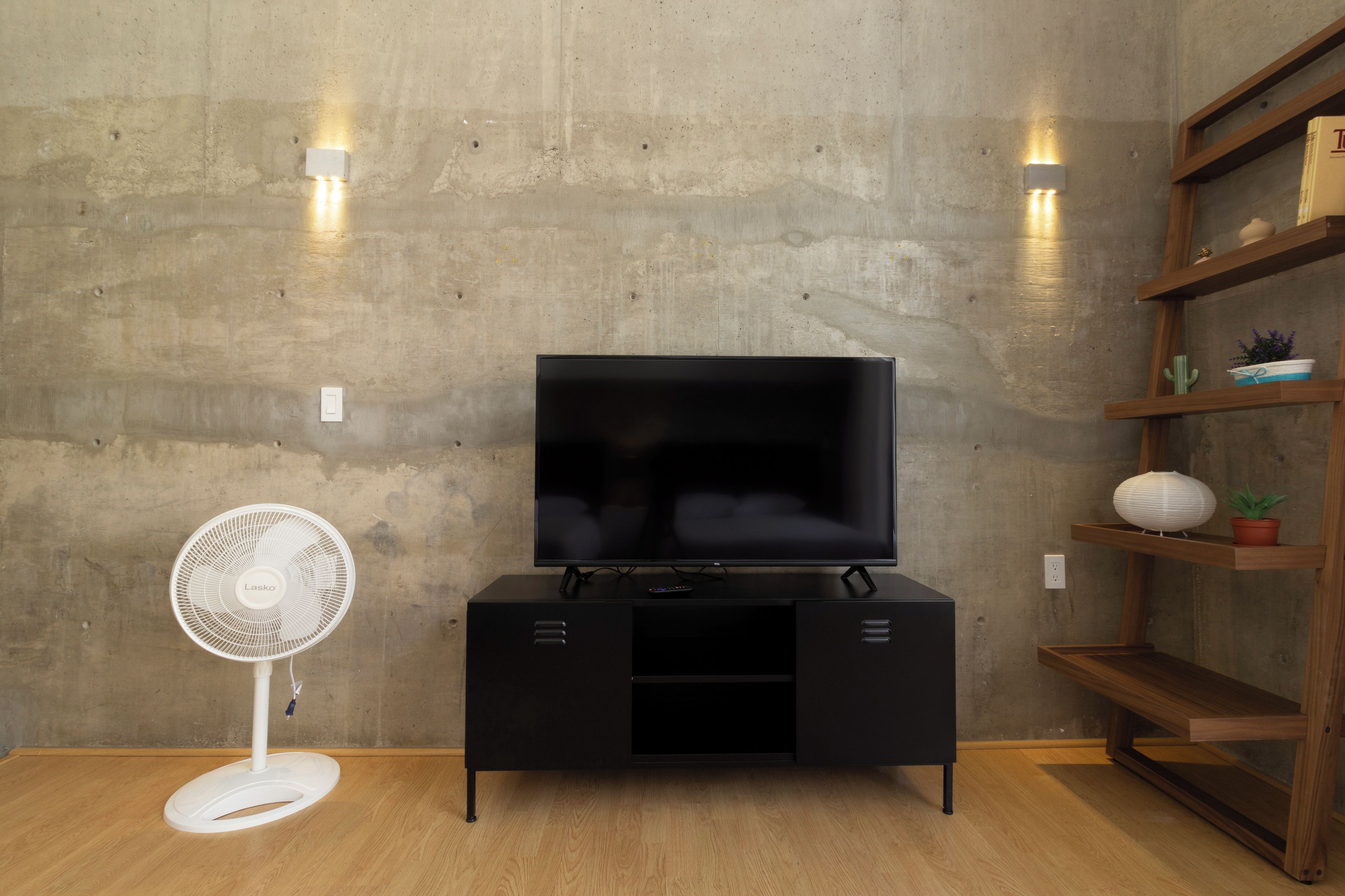 Un salon moderne avec un téléviseur à écran plat posé sur un meuble noir, un ventilateur blanc et une bibliothèque en bois agrémentée d’objets décoratifs.