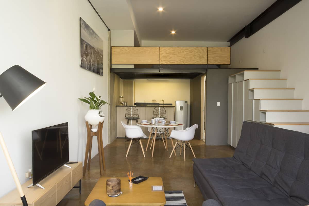 Uma sala de estar moderna com televisão de tela plana, sofá, mesa de jantar com cadeiras e cozinha ao fundo.