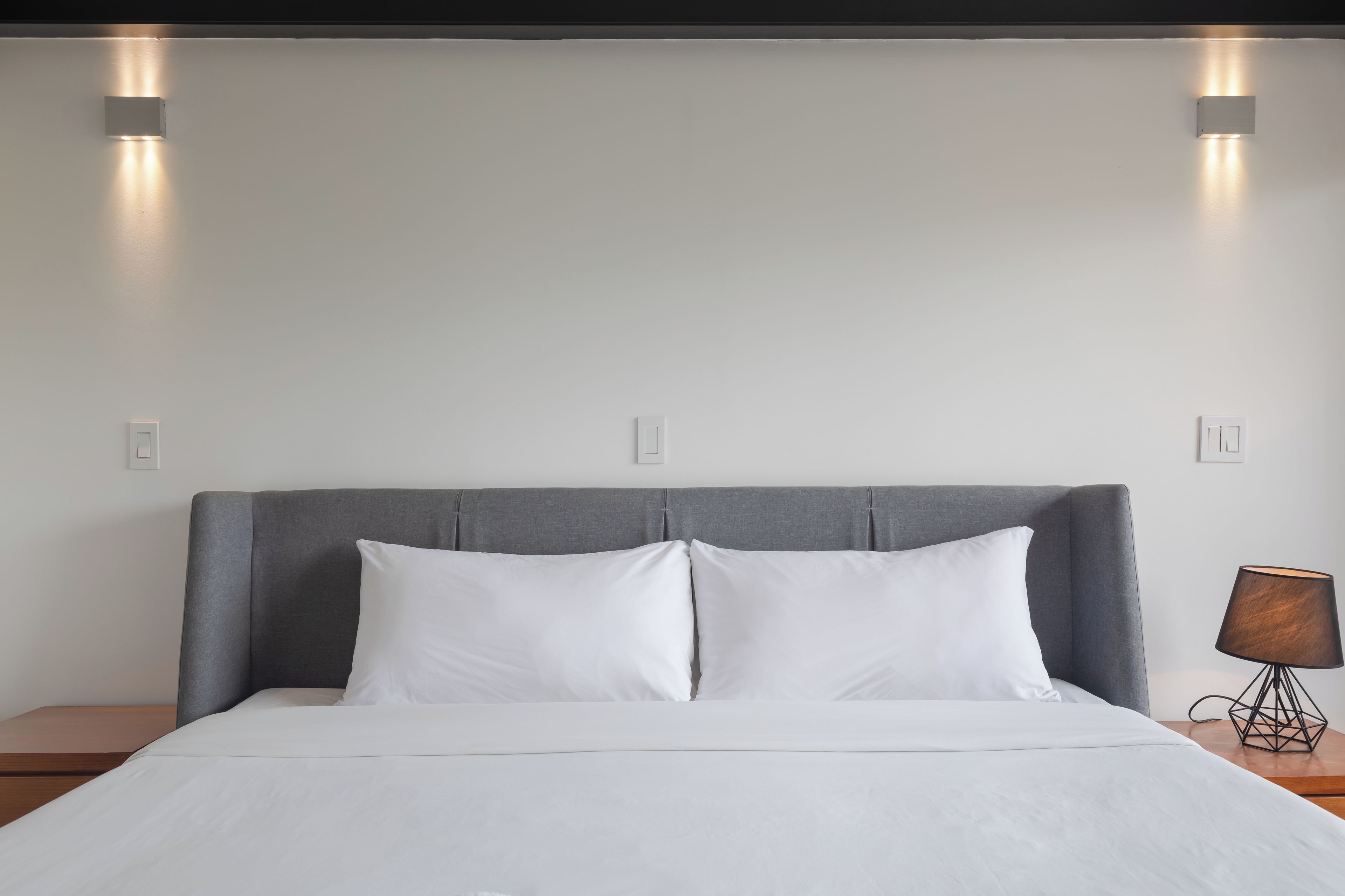 Una cama con cabecera gris, ropa de cama blanca y dos lámparas fijadas a la pared.