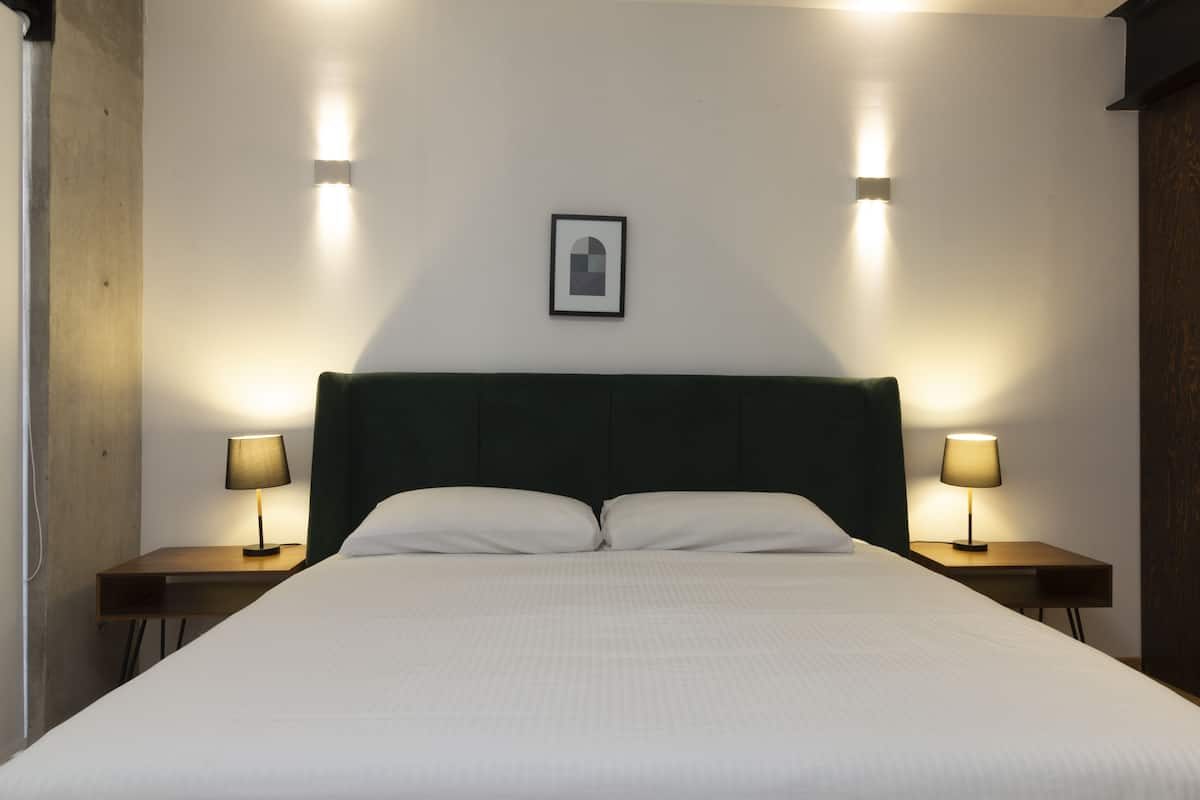 Uma cama bem arrumada com lençóis brancos, duas mesas de cabeceira com abajures e um quadro na parede.
